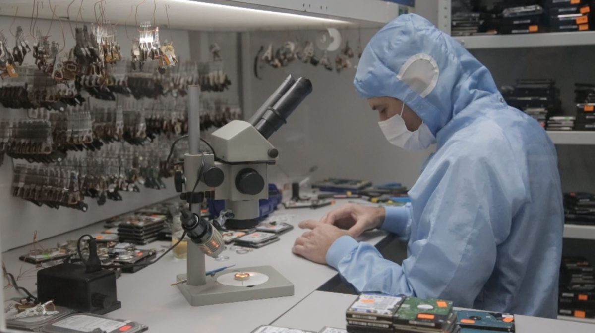 Technicien en combinaison de salle blanche examinant un disque dur au microscope, devant un stock de blocs de têtes de lecture donneurs.